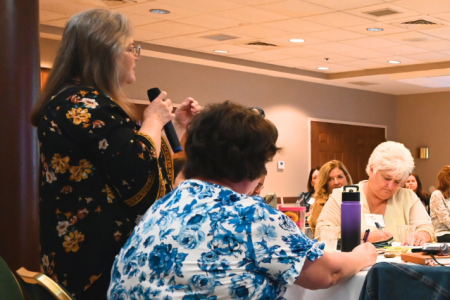 People sitting around a table while a person with a microphone speaks