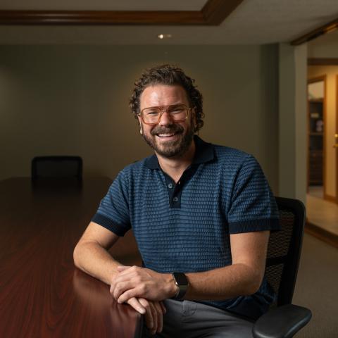 A man sitting at a conference desk with his hands on the desk. He's smiling and wearing a navy and blue polo, glasses, and has curly hair.