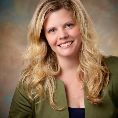 Headshot of Brienne Hooker, a smiling woman with long wavy blonde hair, wearing a green blazer. She is posed in front of a soft, studio-style background.